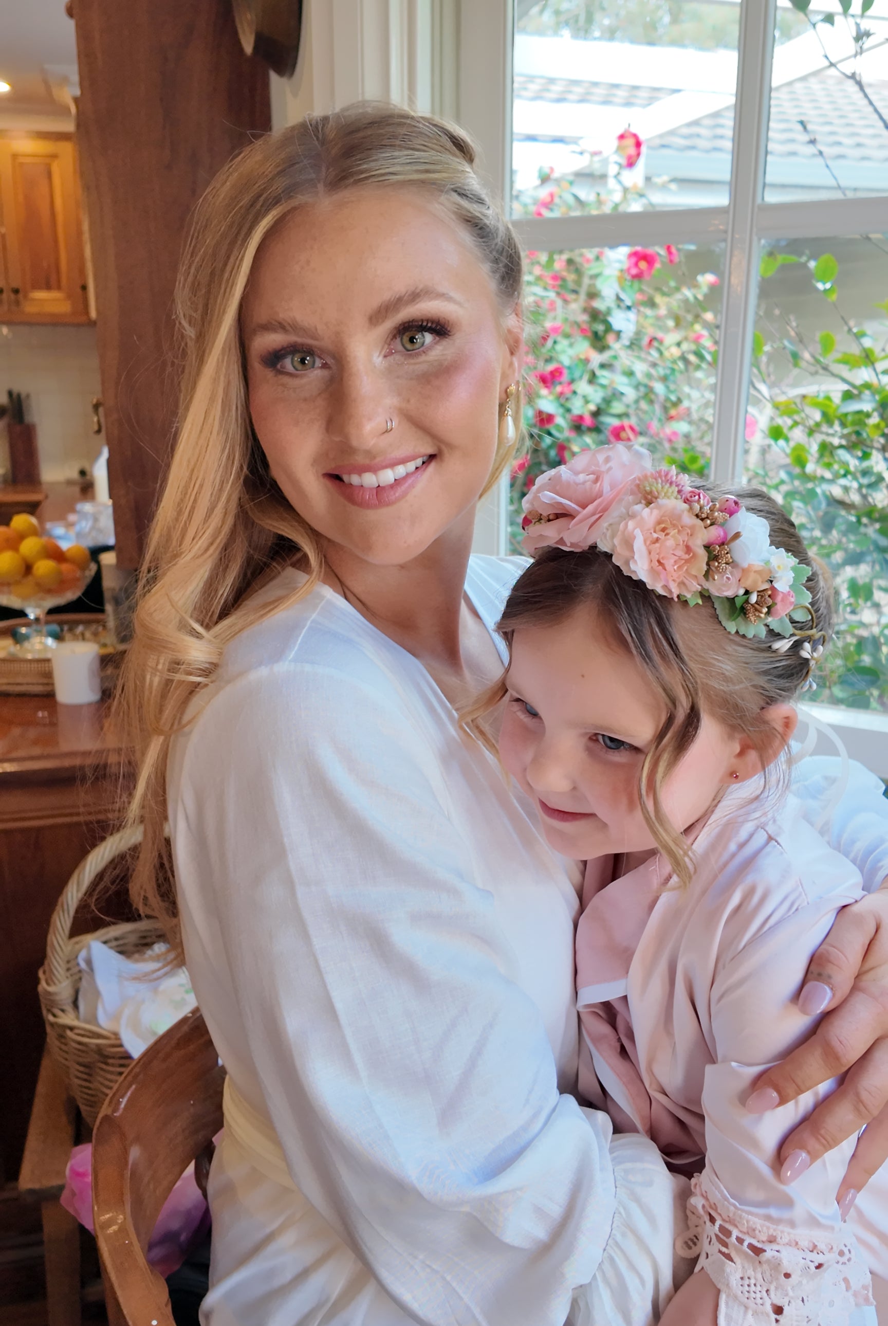 Woman in a white robe holding a young girl with floral hair accessories in a home setting.