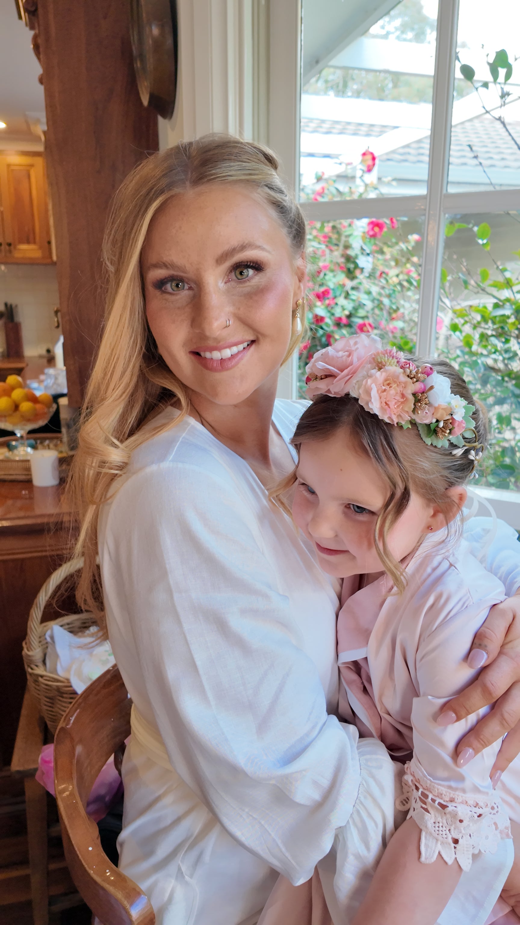 Woman in a white robe holding a young girl with floral hair accessories in a home setting.