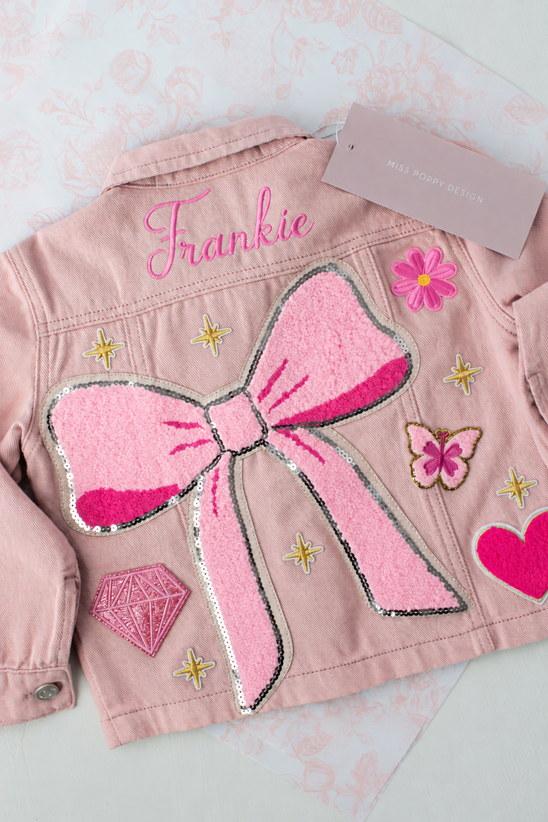 A pink denim jacket for kids, shown from the back, featuring a large pink sequined bow applique, embroidered pink script name 'Frankie', a pink diamond patch, a butterfly patch, a pink heart patch, and gold star patches. A brand tag for 'Miss Rosie Designer' is attached to the collar.