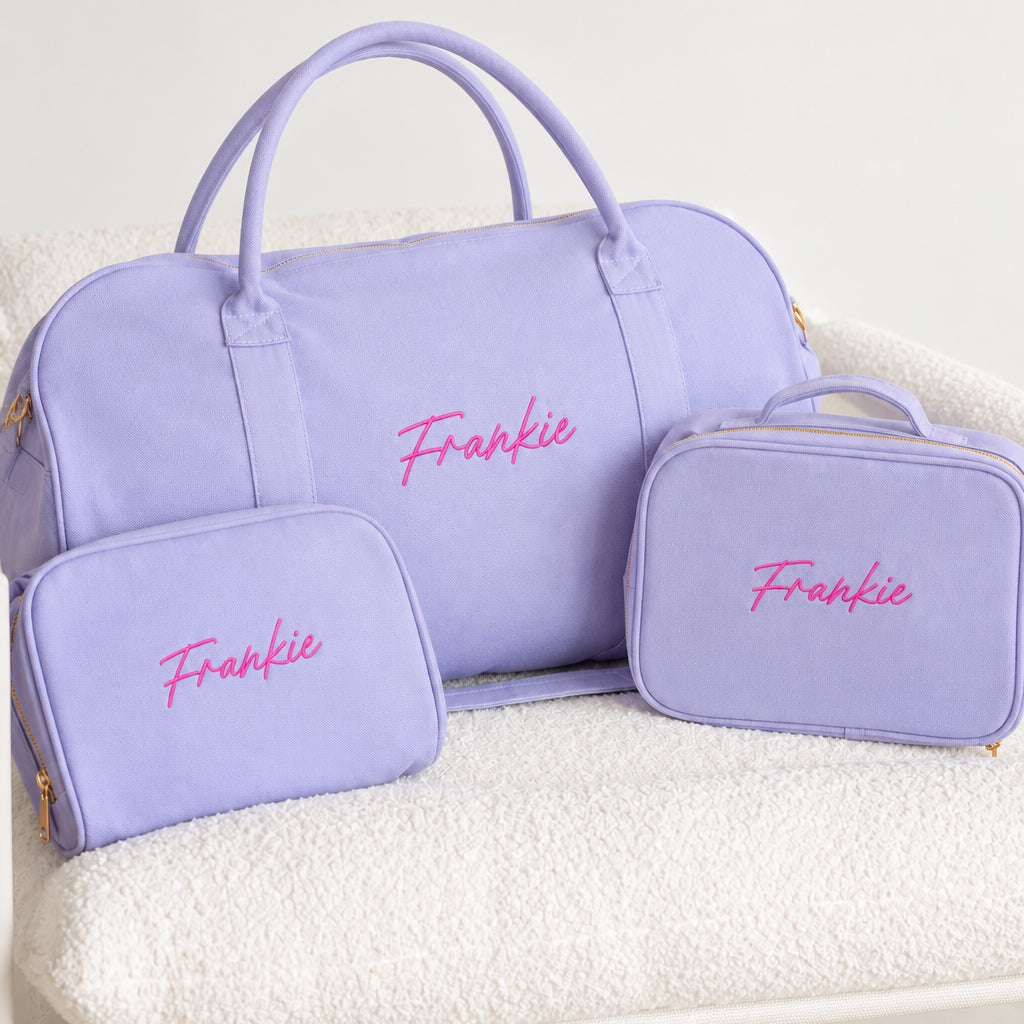 A lavender purple personalized travel set including a duffel bag, a small zip pouch, and a larger rectangular case, all with the name 'Frankie' embroidered in pink on the front, displayed on a white textured blanket.