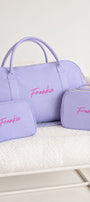 A lavender purple personalized travel set including a duffel bag, a small zip pouch, and a larger rectangular case, all with the name 'Frankie' embroidered in pink on the front, displayed on a white textured blanket.