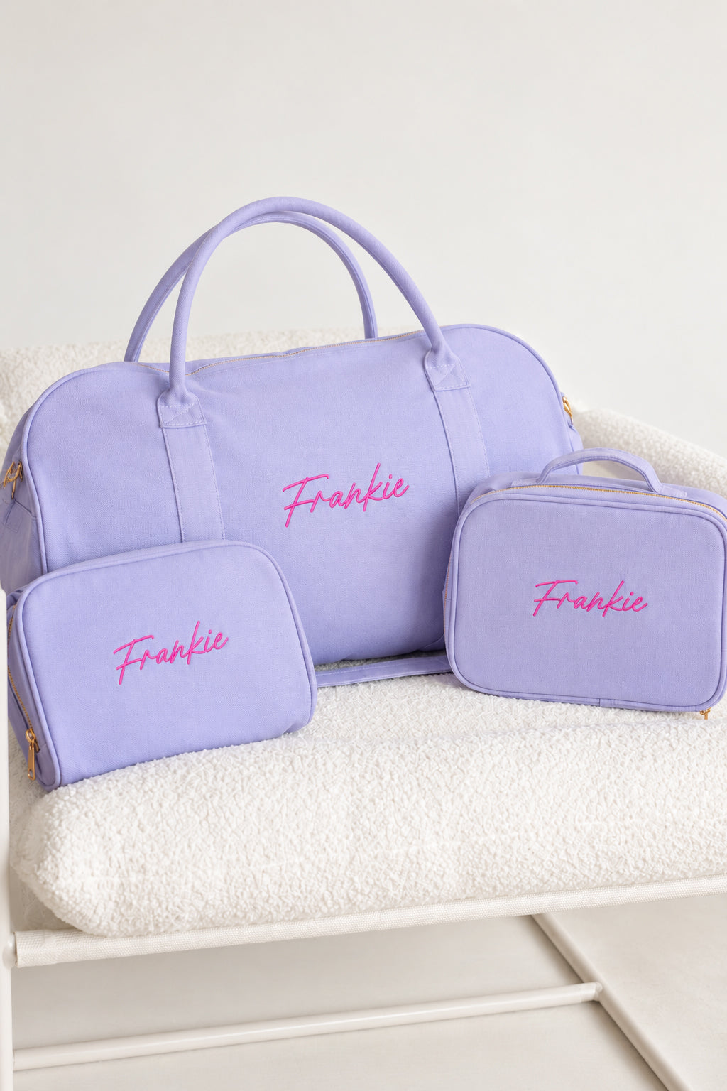 A lavender purple personalized travel set including a duffel bag, a small zip pouch, and a larger rectangular case, all with the name 'Frankie' embroidered in pink on the front, displayed on a white textured blanket.
